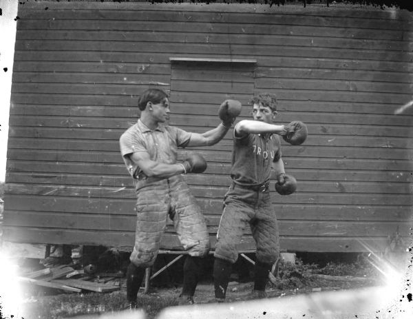 Outdoor portrait of two men wearing baseball uniforms and boxing gloves, posing as if  fighting in front of the wall of a wooden building. Identified as baseball players from Trout Run.
