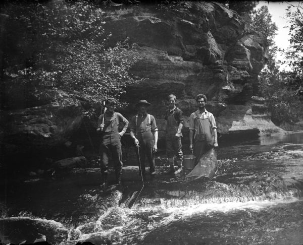 Outdoor group portrait of four men posing standing in ankle-deep water in a river in front of an exposed cliff. One of the men has an axe over his shoulder, and another man is holding a pail and what may be a net. Probably seining for minnows.