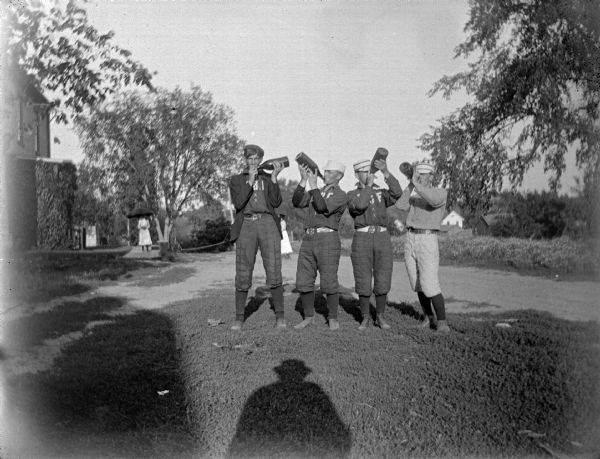 Outdoor group portrait of four men wearing baseball uniforms standing and drinking from large bottles. A woman is in the distance standing and holding an umbrella near a building.