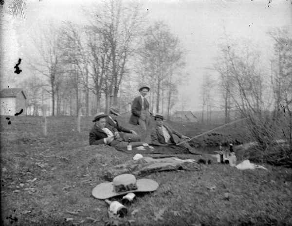 Outdoor group portrait of a four men sitting and lounging on the ground,  and one man standing on a bank near a stream. One of the men is holding a fishing pole. Bottles, hats, and other items are lying in the foreground.