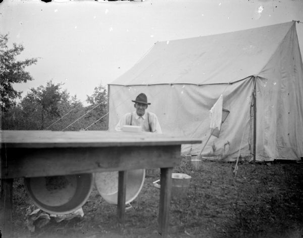 Outdoor portrait across a wooden table with wash bins towards a man sitting in front of a tent at a campsite.