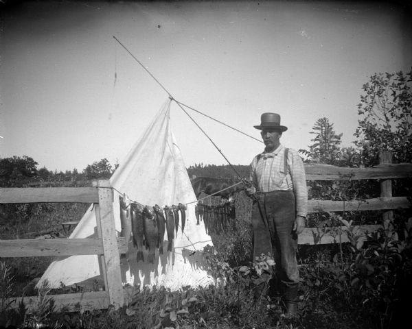 Outdoor portrait of a man standing and holding a fishing pole next to a stringer of fish in the gate of a fence. Behind the fish is a piece of white cloth on a pole for a backdrop. In the background is a horse.