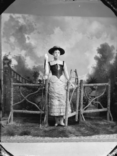 Studio portrait in front of a painted backdrop of a young woman posing standing on fake grass and holding on to a prop gate on a fence. She is holding a Shepherd's crook, and is wearing a hat, and a light-colored dress with a dark-colored bodice. Possibly a theatrical costume.