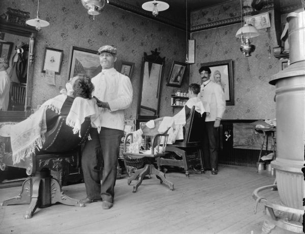 Interior portrait of a barbershop. Two barbers are standing next to two men, each sitting in a barber's chair. Framed prints and mirrors decorate the walls. The side of a woodburning stove in is the right foreground. Identified as the barbershop of Hendricks in Black River Falls.