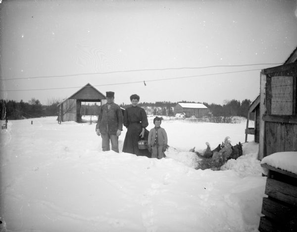 Outdoor group portrait of a man, woman, and a child standing behind a snowbank. On the right is a group of chickens near a wooden building. Farm buildings are in the background