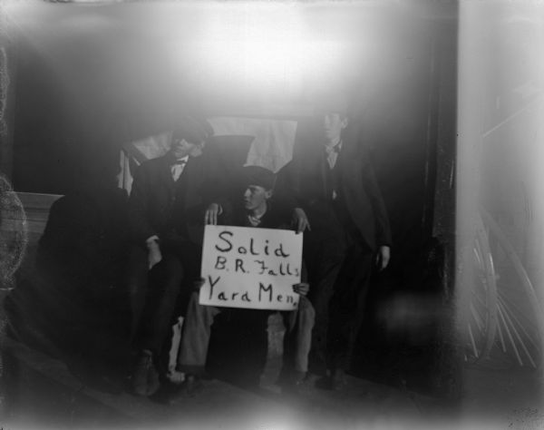 Outdoor portrait of three young men posing standing and sitting. One of the men is holding a sign that reads: "Solid B. R. Falls Yard Men. "
