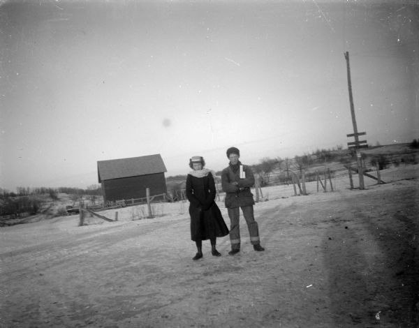 Outdoor portrait of a man and woman posing standing on snow-covered ground. They are bundled in a winter clothing. There is a fence and farm buildings in the background.