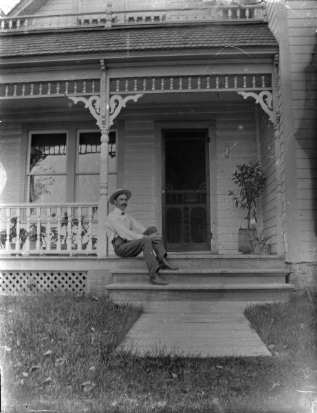 View from yard towards a man posing sitting on the steps of a porch of a wooden house.