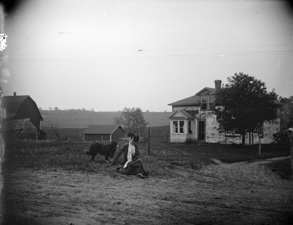 View across road towards a woman sitting on the ground next to a dog in the yard of a wooden house.
