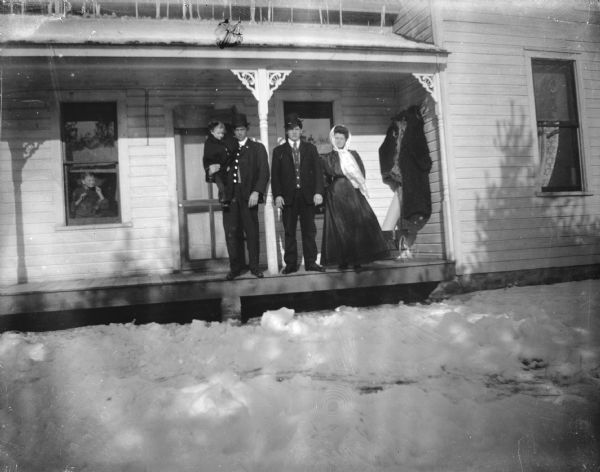 Outdoor group portrait of two men, one holding a child, and a woman posing standing on the porch of a wooden house. The yard is snow-covered. A child inside the house is looking out from a window on the left.