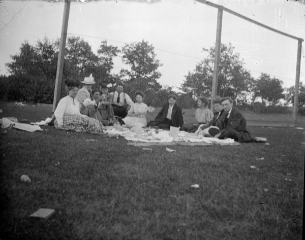 Outdoor group portrait of people posing sitting on the ground at a picnic in front of a tall wire fence. Possibly a baseball backstop.