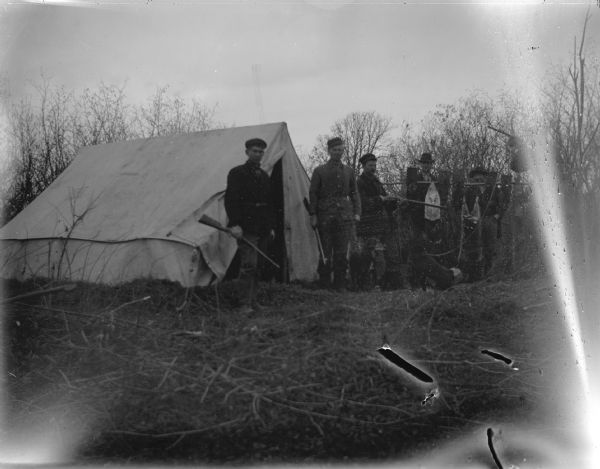 Outdoor group portrait of five men posing standing in front of a tent holding guns. A raccoon is hanging in the background.