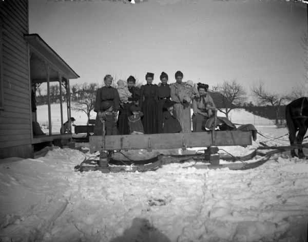 Outdoor group portrait of women and children posing standing on a sled hitched to a single horse. There is a dog on the porch of a a wooden building on the left.