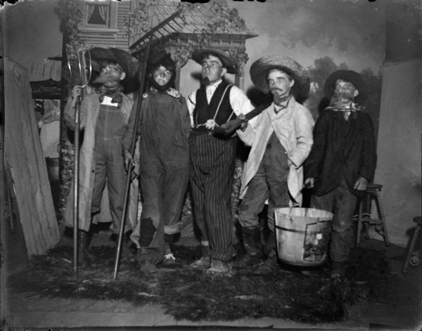 Studio portrait of five children posing standing together on a stage with a painted backdrop. They are wearing theatrical costumes and holding farming tools.