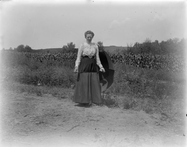 Outdoor portrait of a woman posing standing and holding an umbrella by her side. In the background is a field of corn.