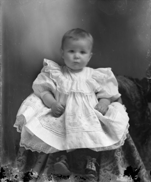 Studio portrait of infant posing sitting in a chair covered with a cloth. The infant is wearing a light-colored dress.
