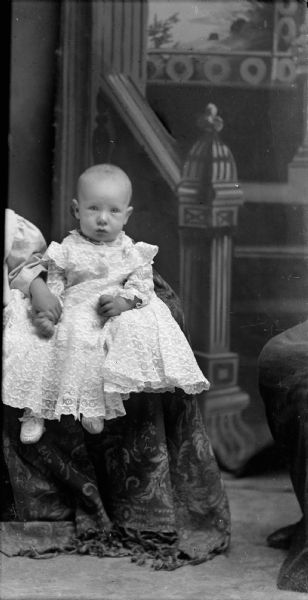 Studio portrait of an infant posing sitting and wearing a light-colored dress, with a child partially visible on the left.