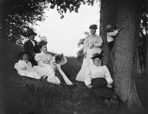 Outdoor group portrait of a man, two women, and a girl sitting, a woman standing, and a boy climbing a tree on a hillside.