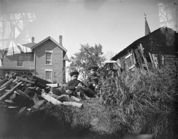 Outdoor portrait of two boys and a girl sitting on the ground next to a pile of wood, with a brick building in the background.