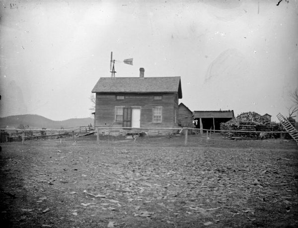 View across empty yard towards a small wooden building with a windmill and outbuildings in a dirt yard.
