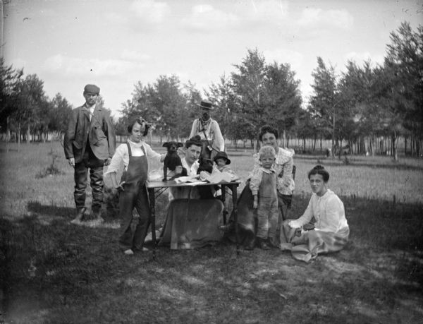 Outdoor group portrait of men, women, and children posing standing and sitting around and near a small table with two dogs on top of it.