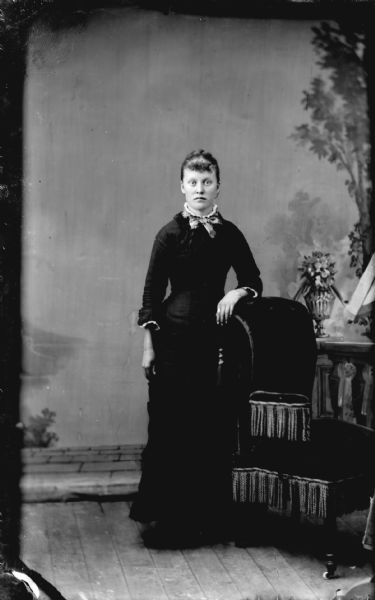 Studio portrait of a woman standing with her left arm resting on a chair on the right.