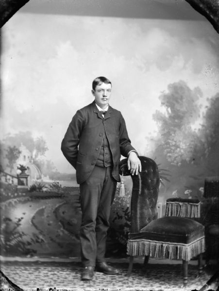 Studio portrait of man posing standing with his left hand resting on a chair on the right. He is wearing a dark-colored suit coat, vest, and trousers.