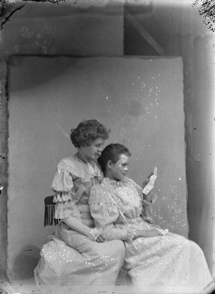 Studio portrait of two women posing sitting in a chair, looking at a letter held by the woman sitting lower on the right. Both women are wearing light-colored dresses.