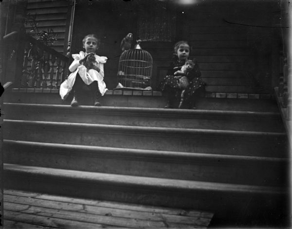 Outdoor view of two girls posing sitting on the top of porch steps, on either side of a bird cage with a parrot on top of it. Both girls are holding cats in their laps.