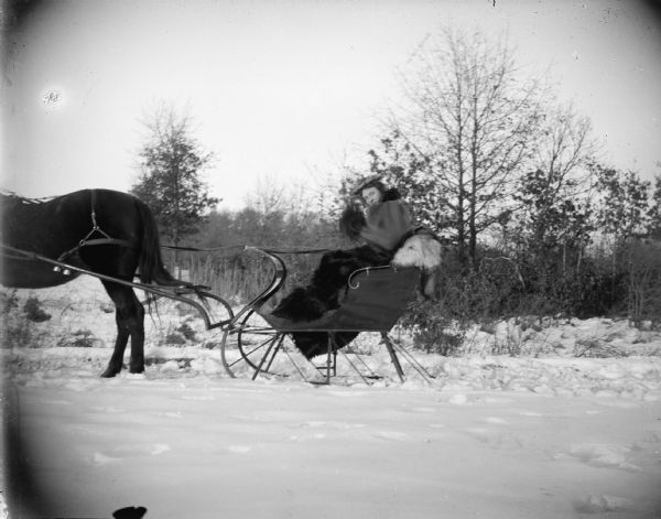 Outdoor view of a woman posing sitting in a sleigh pulled by a horse on a snow-covered road.