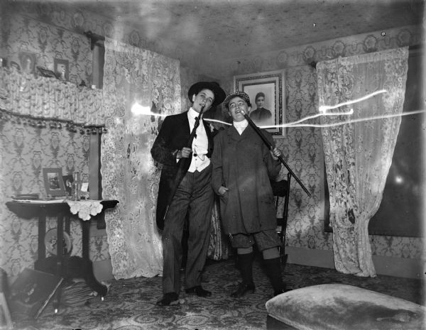 Indoor view of two women dressed in men's clothing posing standing and holding an umbrella and walking stick.