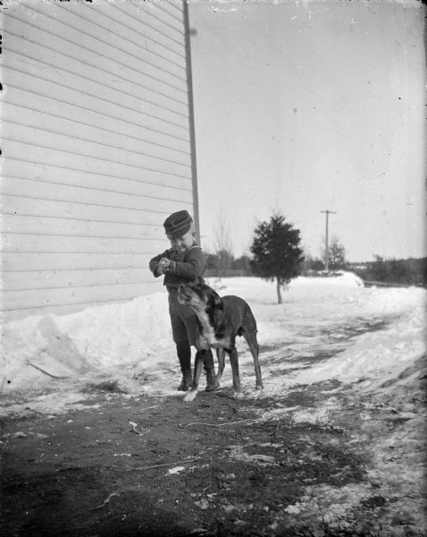 Outdoor view of a small boy holding a gun posing standing on a road between piles of snow and next to a dog.