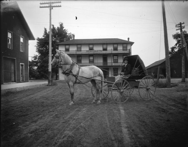 Outdoor view of a man posing sitting in a buggy pulled by a horse on a town street. Location identified as in front of the Merchants Hotel.