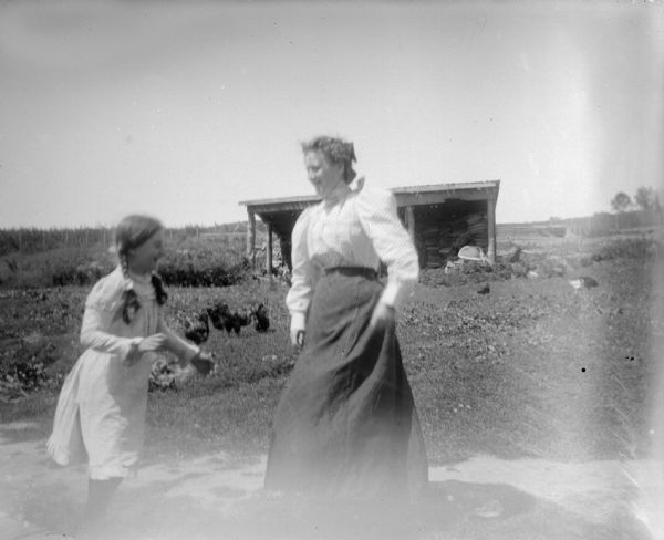 Outdoor view of a woman and a girl posing standing in a yard. There are chickens and a wooden structure in the distance.