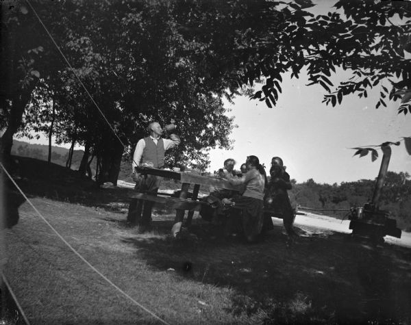 Outdoor view of a man standing and drinking from a bottle next to a picnic table. Women and girls are sitting are sitting at the picnic table, and one of the women has her arm around a dog that is standing on its hind legs with its paws on the bench. On the right is a woodstove with pots and kettles. There are ropes in the left foreground, and one rope is tied to the side of the picnic table.