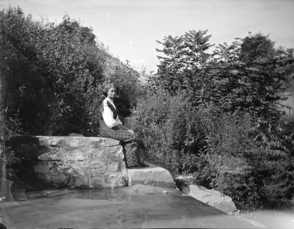 Outdoor view of a woman posing sitting on a stone wall. She is wearing a dark-colored dress.