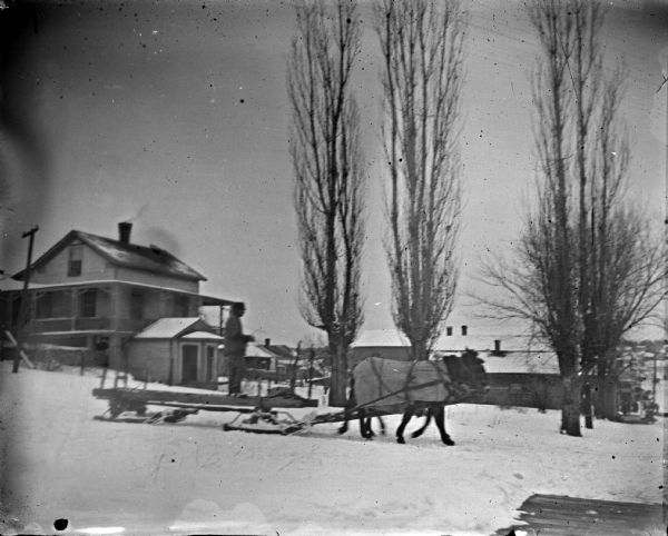 Outdoor view of a man posing standing on a sled pulled by a team of two horses on a snow-covered road.