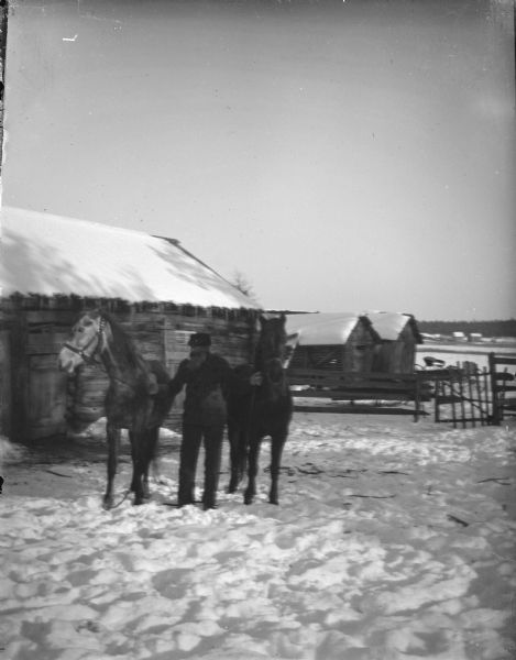 Outdoor view of a man posing standing and displaying two horses on a snow-covered ground in front of several wooden buildings.