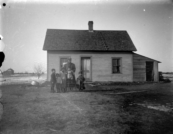 Outdoor group portrait of a man and five children posing standing in the yard of a small wooden house. Possibly Native American.