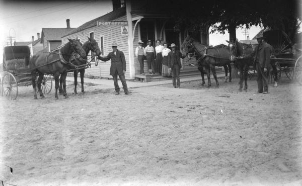 Outdoor group portrait of two men displaying two teams of two horses which are hitched to buggies in front of a post office building. A group of people are standing on the porch of the post office.