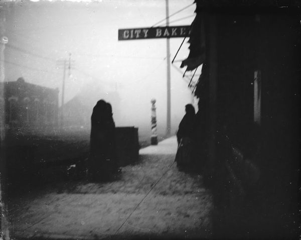Outdoor view of women standing on a wooden walkway on a foggy town street. Location identified as the walkway in front of the City Bakery.