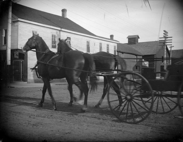 Outdoor view of a team of two horses pulling a wagon on a town street. Location identified as the intersection of Main and First Street looking south.