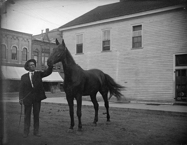 Outdoor view of a man displaying a horse on a town street. Location identified as South First street looking northeast toward the intersection of Main and First Streets.