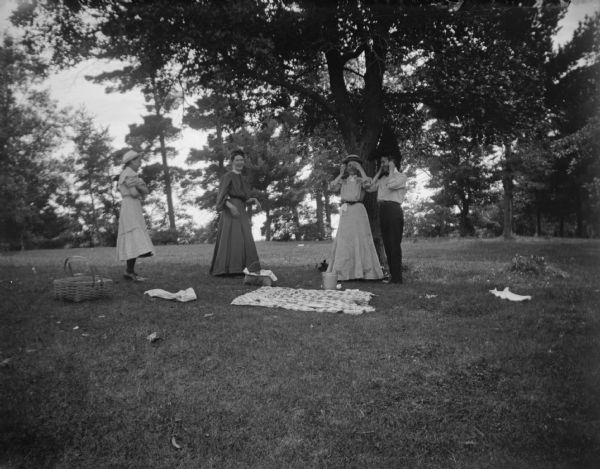 Outdoor group portrait of three women and a man posing standing near a picnic in front of and among several trees.