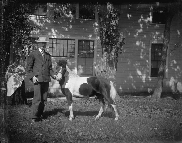 Outdoor view of a man posing standing and displaying a small pony in front of a woman and a wooden house.