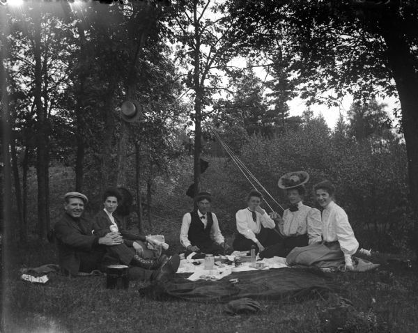 Outdoor view of men and women posing sitting at a picnic. Their hats are hanging on the trees.
