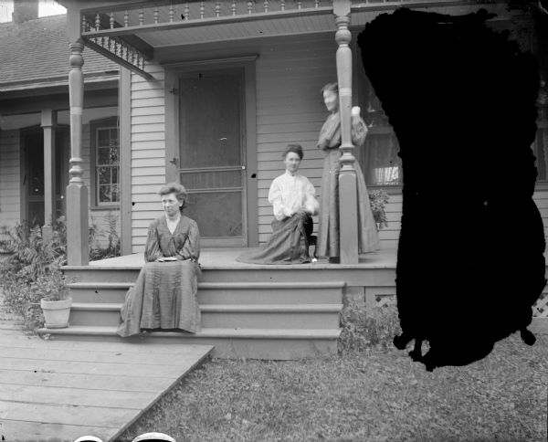 Outdoor view of two women posing sitting, and a woman posing standing on the porch of a wooden house.