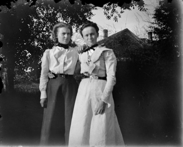 Outdoor view of two women posing standing in front of trees and a house in the distance.
