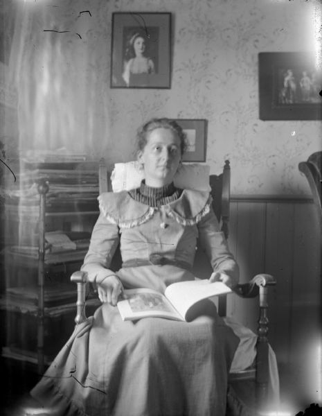 Indoor view of a woman posing sitting in a chair and holding a book.