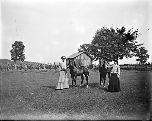 Outdoor view of two European American women posing standing and displaying two horses in a field. There is a wooden building in the background.
